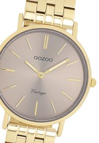 Montre en métal doré avec un cadran rond, un cadran dégradé brun clair, des index horaires dorés et un bracelet à maillons. Présente le texte "OOZOO" et "Vintage".