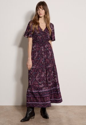 Frau trägt ein dunkellila Maxikleid mit Paisley-Muster, flatternden Ärmeln und schwarzen Knöchelstiefeln, steht vor einer schlichten Wand.