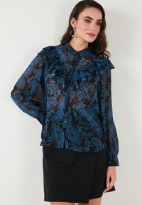 Sheer blue paisley blouse with ruffled front, long sleeves, and a button closure, paired with a black skirt. Smooth texture.