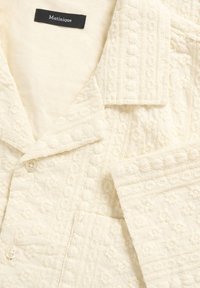 Veste de couleur crème avec motif floral brodé, deux boutons et une étiquette noire portant l'inscription "Matinique" à l'intérieur du col.