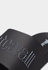 Gros plan sur une sandale noire à bride avec le nom de la marque "Cavalli" en relief sur la lanière, sur un fond blanc.