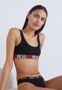 Soutien-gorge de sport noir avec une large bande élastique arborant le texte et le logo "DIESEL" en blanc et rouge, associé à un string noir assorti. Texture lisse.