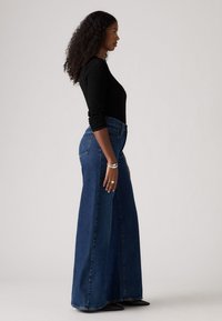 Jean en denim large en bleu foncé avec une taille haute, dotés de poches avant, de surpiqûres sur les poches arrière et d'une texture lisse.