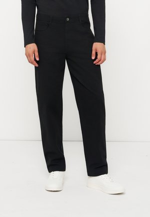 Personne portant un pantalon noir droit, un haut noir à manches longues et des baskets blanches, debout devant un fond blanc uni.