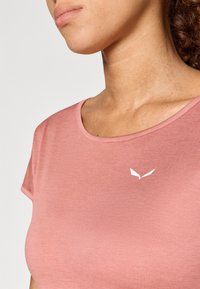 Gaiši rozā t-krekls ar īsām piedurknēm un noapaļotu kakla izgriezumu, ar mazu baltu logotipu uz krūts, izgatavots no mīkstas, gludas auduma.