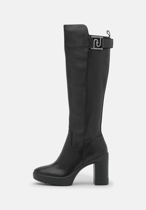 Botte en cuir noire montant jusqu'au genou avec talon bloc, bout arrondi, semelle plateforme et une sangle décorative avec boucle métallique près du haut.