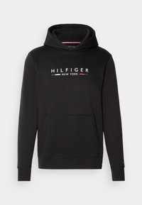 Svart hoodie i mjuk tyg, med en kenguruficka och ribbade muddar. Framsidan visar texten "HILFIGER NEW YORK" i vitt, rött och blått.