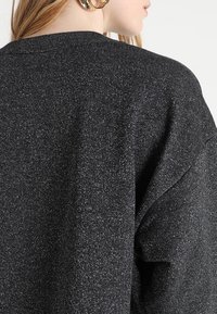 Suéter negro de punto con un corte holgado, que presenta una tela texturizada y brillante, con hombros caídos, mostrando parte de un gran pendiente.