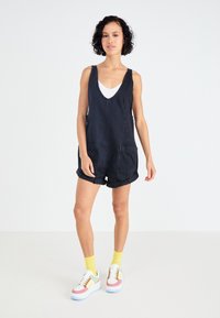 Salopette en denim noir avec un décolleté en V profond, deux poches avant et des shorts roulés. Assortie avec un débardeur blanc et des chaussures de sport colorées.