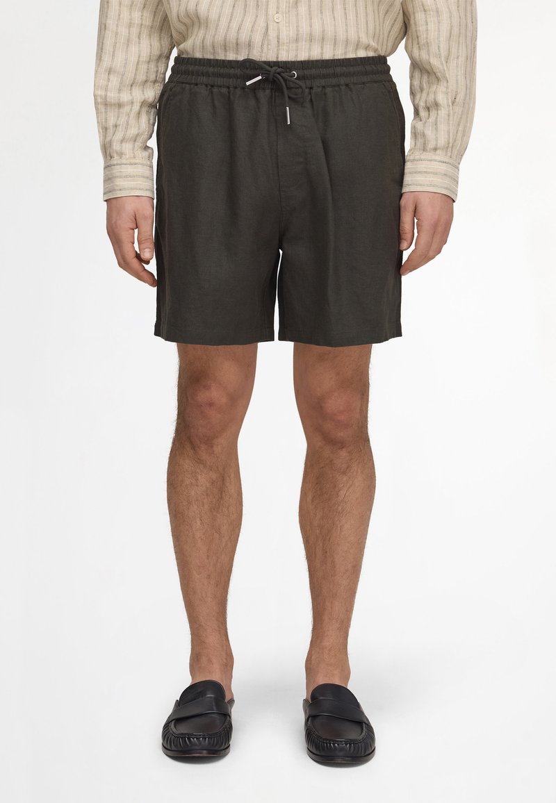 Homme portant un short noir à cordon, une chemise beige rayée à manches longues et des chaussures noires sans lacets, debout droit.