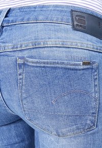 Jeans de mezclilla azul con textura desgastada. Cuenta con un bolsillo trasero con una línea curva cosida y una etiqueta de marca en la cintura de material oscuro.