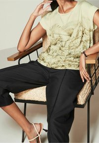 Lys grøn bluse med korte ærmer og blomstermotiv samt bladværk, parret med sorte bukser med brede ben. Modellen sidder i en vævet stol iført beige sandaler.