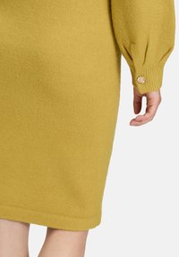 Vestito in maglia di un giallo senape con una finitura texturizzata, caratterizzato da maniche a palloncino e un bottone floreale decorativo vicino al polsino.