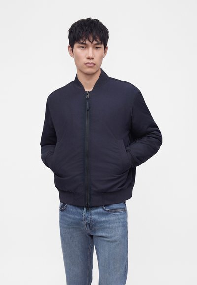 Jeune homme les mains dans les poches, portant une veste bomber zippée bleu marine foncé et un jean bleu clair sur un fond blanc uni.
