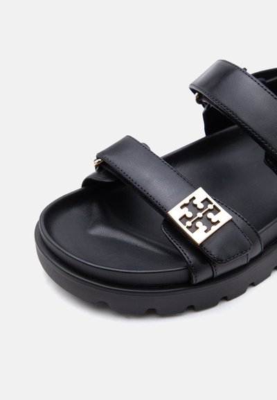 Μαύρο δερμάτινο σανδάλι με δύο ρυθμιζόμενα λουράκια και ένα χρυσό διακοσμητικό τετράγωνο έμβλημα στο μπροστινό λουράκι, μαξιλαρωμένη σόλα και υφή από καουτσούκ στην εξωτερική σόλα.