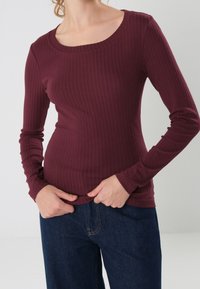 Burgundy long-sleeve ribbed top with a scoop neckline; fitted design and stretchy texture. Paired with dark denim pants.