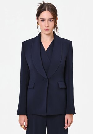 Giovane donna con capelli castani legati, che indossa orecchini dorati e un blazer blu scuro su misura con pantaloni abbinati su sfondo bianco.
