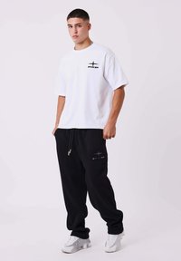 Giovane uomo in piedi che indossa una maglietta bianca PXP, pantaloni sportivi neri PXP e sneakers bianche su uno sfondo bianco.
