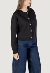 Felpa nera con girocollo, bottoni frontali e maniche lunghe. Realizzata in materiale morbido con vita aderente. Indossata con jeans a gamba larga blu.