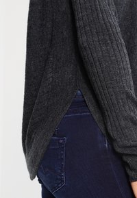 Dunkelgrauer, gerippter Strickpullover mit seitlichem Schlitz, getragen über dunkelblauer Denim-Jeans, hebt Stoffstrukturen und Passform hervor.