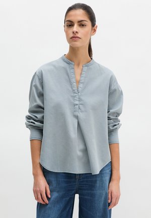 Femme aux cheveux foncés attachés, portant un chemisier bleu clair à manches longues avec une encolure fendue et un jean bleu, sur un fond uni.