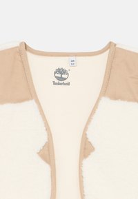 Gilet en polaire beige avec des panneaux en fausse fourrure crème. Il présente un col rond et le logo Timberland au centre. Texture douce avec un design décontracté.