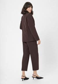 Bruin op maat gemaakt pak met een lange blazer en verkorte broeken. Glad stof, eenvoudig ontwerp en gecombineerd met zwarte hakken.