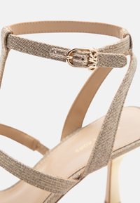 Σανδάλι με ψηλό τακούνι και λουράκια από λαμπερό χρυσό ύφασμα, κλείσιμο στον αστράγαλο με αγκράφα που διαθέτει μικρό μεταλλικό λογότυπο της μάρκας.