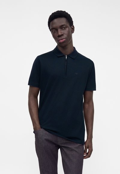 Jeune homme portant un polo noir à manches courtes avec une fermeture éclair à l'avant et un pantalon gris foncé, debout devant un fond blanc uni.