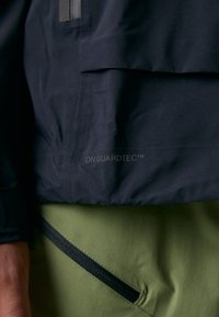 Veste noire en tissu lisse et résistant à l'eau, avec une poche poitrine. Pantalons verts avec un détail de fermeture éclair noir à la taille.
