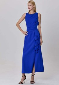 Femme portant une robe midi sans manches bleu roi avec des volants et une fente frontale, associée à des talons hauts noirs à bride de cheville, debout.