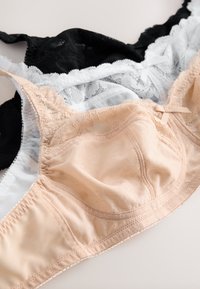 Trois soutiens-gorge en dentelle beige, blanc et noir, superposés sur une surface claire, mettant en valeur un tissu délicat et de petits nœuds à l'avant.