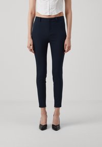 Marinblå skreddersydde bukser med slim fit, laget av elastisk stoff. Har en frontglidelås og små glidelåser på sidene ved anklene.