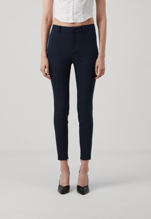 Marinblå skreddersydde bukser med slim fit, laget av elastisk stoff. Har en frontglidelås og små glidelåser på sidene ved anklene.