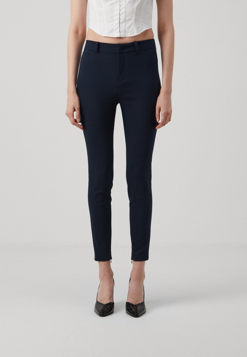 Marinblå skreddersydde bukser med slim fit, laget av elastisk stoff. Har en frontglidelås og små glidelåser på sidene ved anklene.