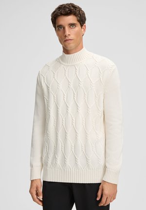 Mann in einem cremefarbenen, strukturierten Pullover mit hohem geripptem Kragen und schwarzen Hosen, der vor einem schlichten, hellgrauen Hintergrund steht.