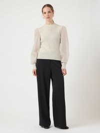 Woman standing wearing beige sweater with sheer puff sleeves, black wide-leg trousers, and black shoes, against plain light background.