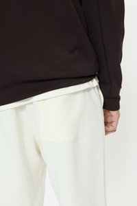 Približen posnetek osebe, ki nosi temno rjavo majico z dolgimi rokavi čez bele hlače z vidnim zadnjim žepom in roko ob strani.