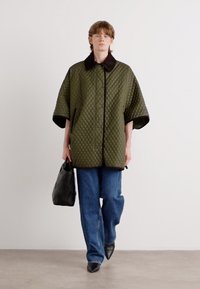 Gequilted groene overshirt met brede mouwen, zwarte kraag en voorzakken; gecombineerd met blauwe jeans en een zwarte, gestructureerde handtas.