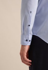 Camicia azzurra chiara con ricamo di una singola rosa sul polsino, due bottoni scuri e tessuto morbido con una vestibilità strutturata.