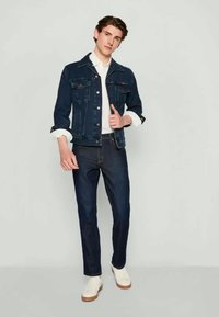 Jaqueta de ganga masculina em azul escuro, com um corte clássico, bolsos frontais e botões metálicos, combinada com jeans escuros e sapatilhas brancas.