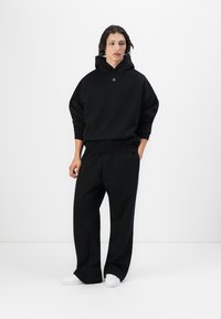 Svart oversized hettegenser med lommer foran og snøring, kombinert med løstsittende svarte joggebukser. Hvite sneakers fullfører antrekket.