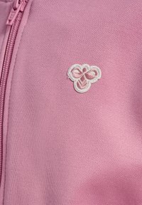 Zbliżenie na różową teksturowaną tkaninę z dopasowanym zamkiem błyskawicznym i małym białym wyhaftowanym abstrakcyjnym logo na wysokości klatki piersiowej.