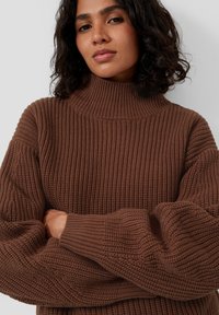 Brązowy sweter z dzianiny z wysokim golfem, prążkowaną fakturą i oversize'owymi rękawami. Materiał wydaje się gruby i ciepły, odpowiedni na chłodniejsze dni.