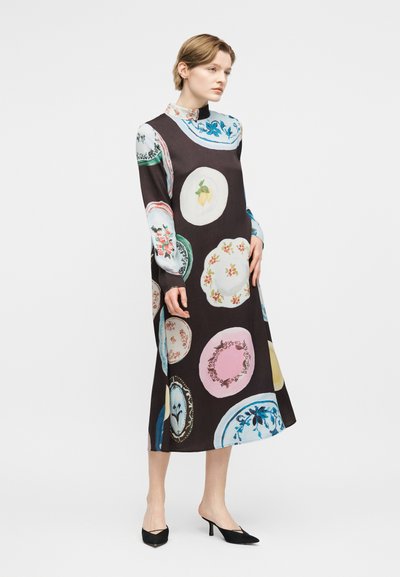 Γυναίκα που στέκεται, φοράει μαύρο midi φόρεμα με μακριά μανίκια και πολύχρωμα σχέδια με λουλούδια και φρούτα, συνδυασμένο με μαύρες μυτερές γόβες.