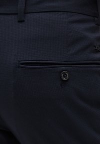 Κοντινό πλάνο τσέπης σε παντελόνι navy blue με υφή, με κουμπί κλεισίματος και λεπτομέρεια θηλιάς για ζώνη.