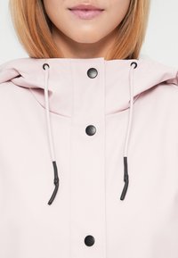 Κοντινό πλάνο ενός ατόμου με ανοιχτόχρωμο δέρμα και ίσια ξανθά μαλλιά που φοράει μια ανοιχτό ροζ ζακέτα με μαύρα κουμπιά και λευκές κορδόνια.