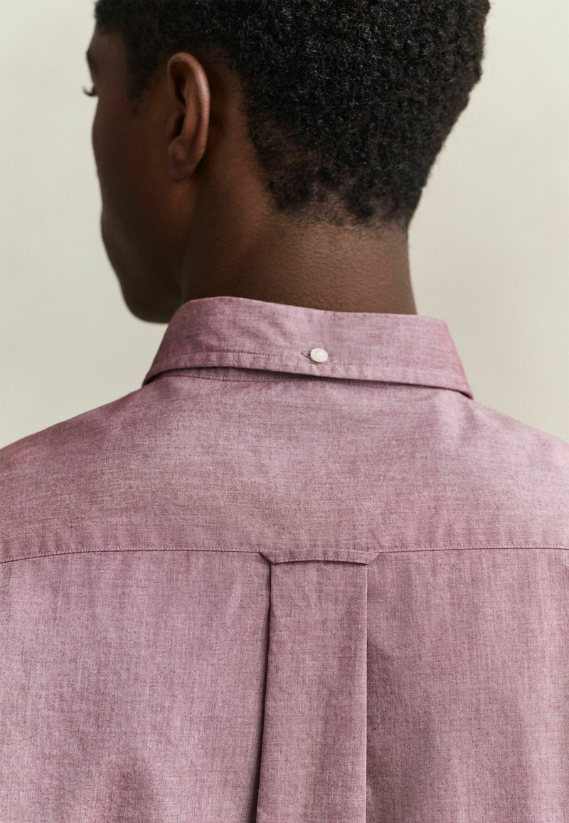 Homme aux cheveux courts et bouclés portant une chemise à col violet clair, vu de dos sur un fond uni.