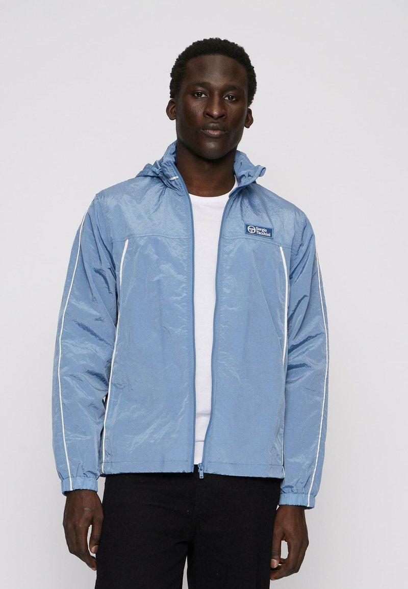 Světle modrá bunda se zipem vpředu, elastickými manžetami a bílými bočními pruhy. Obsahuje kapuci a malé logo na hrudi.