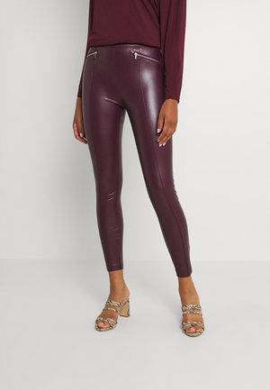Person trägt burgundrote Leggings aus Kunstleder mit Reißverschlusstaschen vorne und beigefarbene High-Heel-Sandalen mit Schlangenledermuster, steht vor einem weißen Hintergrund.